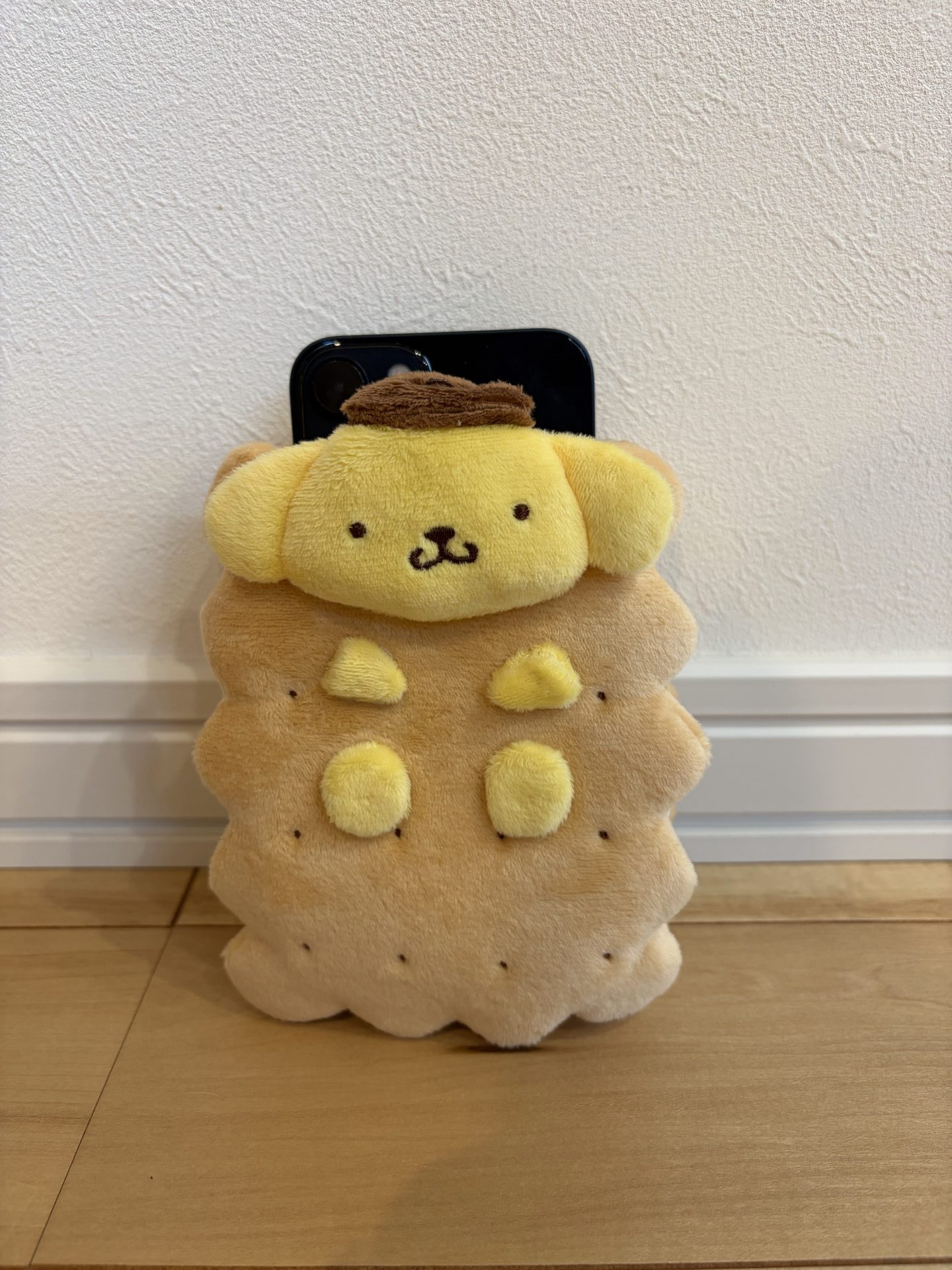 ポムポムプリンがココナッツサブレに挟まったデザインのぬいぐるみポーチ。スマートフォンとを入れてイズ感を比較している写真。